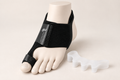 A black toe spacer brace shown on a mannequin foot, with a translucent white silicone toe spacer placed beside it on a light beige background.