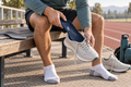A man sitting on a wooden bench at an outdoor track and field wearing a light gray running shoe and putting on a navy blue insole. Another insole and a second running shoe are on the bench beside him.